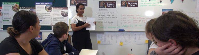 Teacher stands in front of class showing sheet of paper
