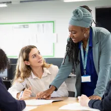 Teacher leaning over desk pointing to sheet. Student looking up at teacher.